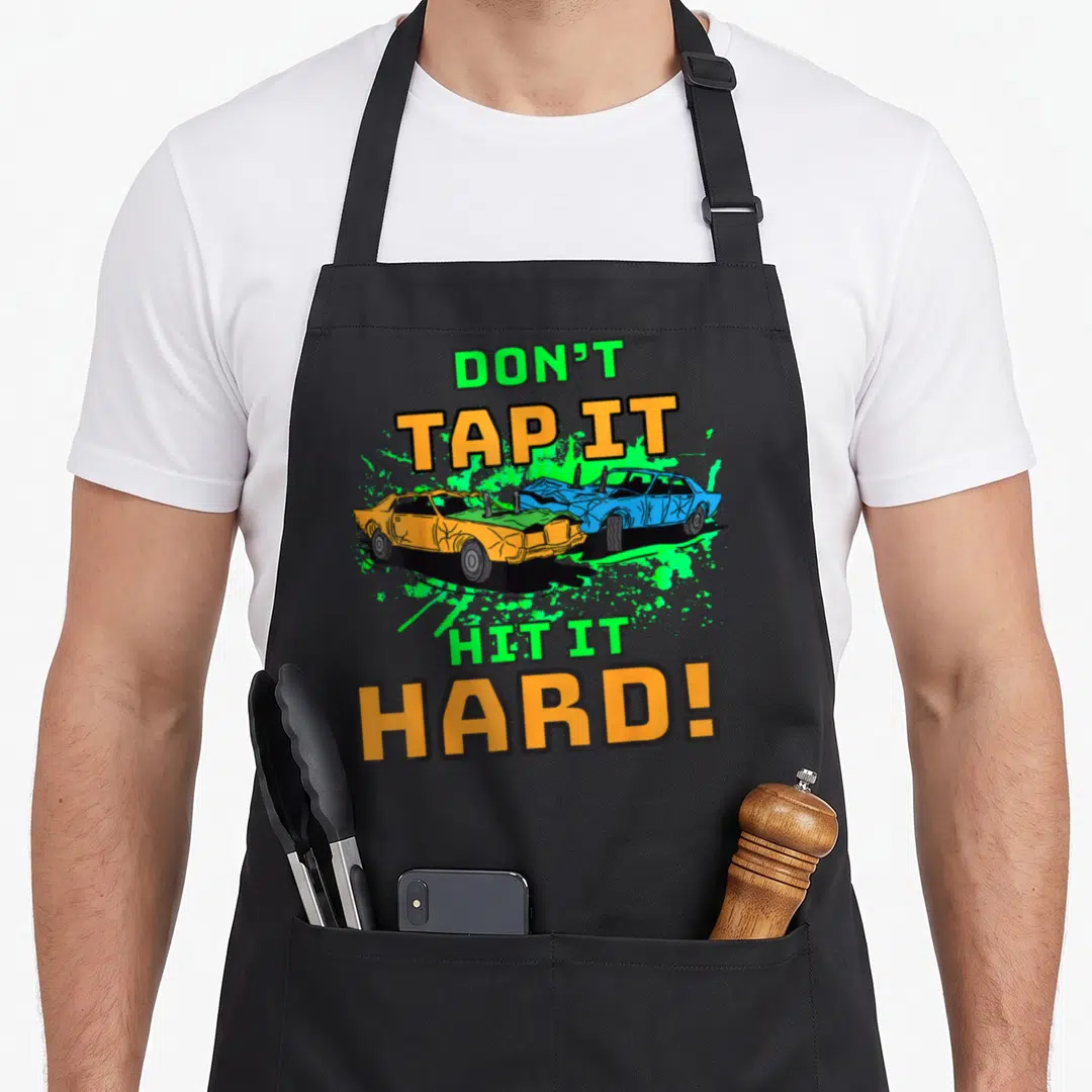 Hit auto apron - Don't Tap It Hit It Hard text surrounding two crashing orange and blue cars on green splatter — shown on a black cotton canvas apron against a white background