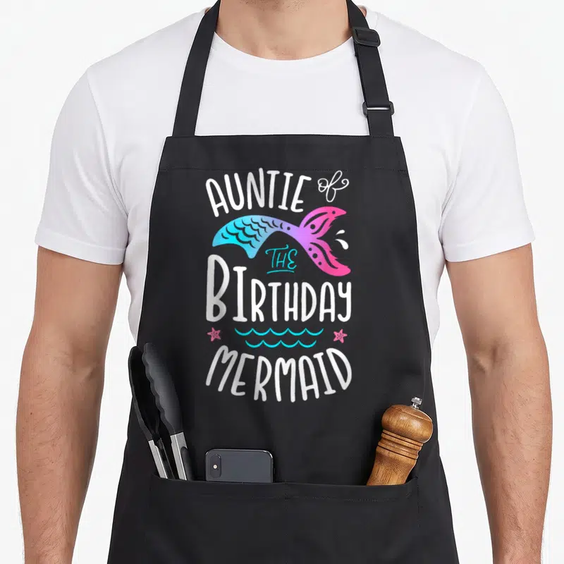 Aunt family aprons - Cartoon mermaid tail with AUNTIE of THE BIRTHDAY MERMAID text and two pink starfish — shown on a black cotton canvas apron against a white background
