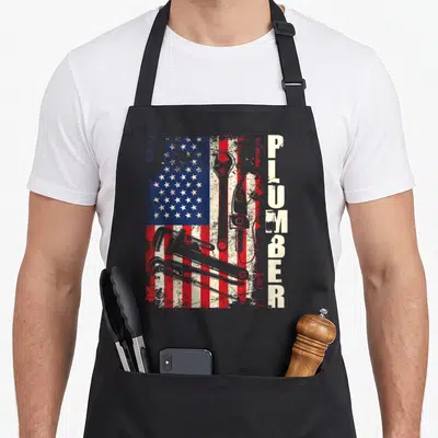Plumber patriotic apron - Distressed US flag with three plumbing tools and vertical PLUMBER text — shown on a black cotton canvas apron against a white background