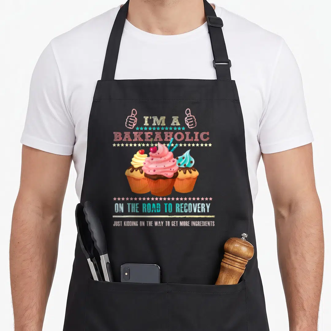 Humour cupcake apron - Three cupcakes with pink frosting and cherries under thumbs up hands and bakeaholic text — shown on a black cotton canvas apron against a white background