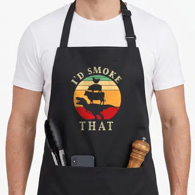 Dinosaur meat aprons - Retro I'd Smoke That text over striped circle with stacked chicken pig cow and T-Rex silhouettes — shown on a black cotton canvas apron against a white background