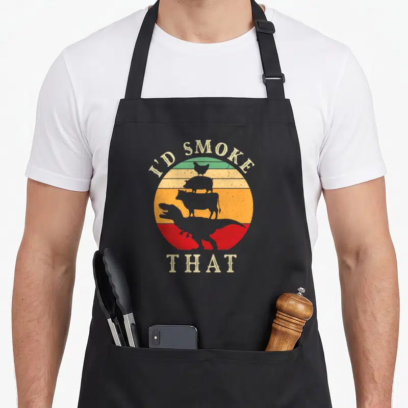 Dinosaur meat aprons - Retro I'd Smoke That text over striped circle with stacked chicken pig cow and T-Rex silhouettes — shown on a black cotton canvas apron against a white background