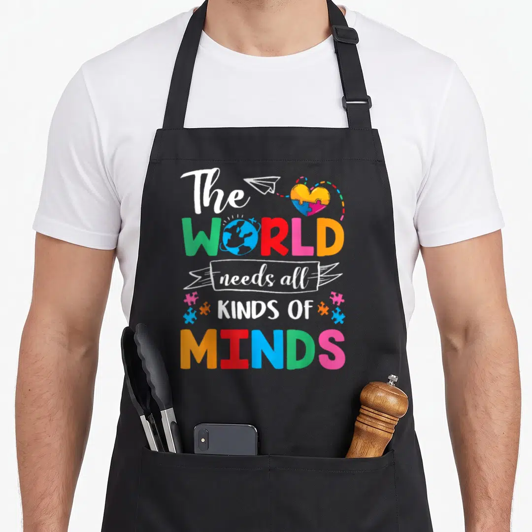 Autism novelty apron - Colorful text The World Needs All Kinds of Minds with puzzle pieces and earth graphic — shown on a black cotton canvas apron against a white background