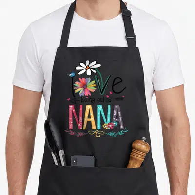 Called nana aprons - Love being called NANA text with daisy, bird, arrows, and patterned colorful letters — shown on a black cotton canvas apron against a white background