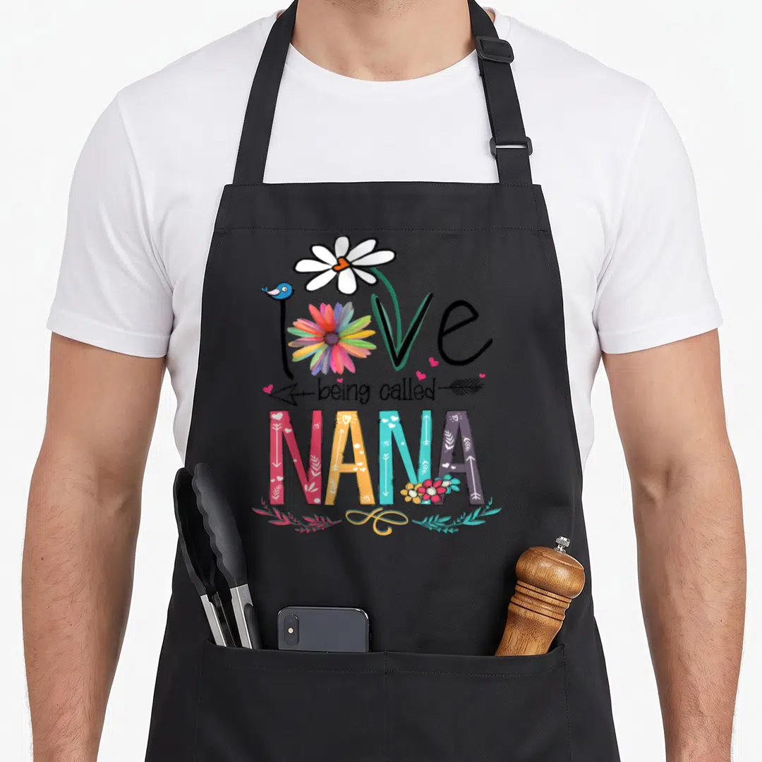 Called nana aprons - Love being called NANA text with daisy, bird, arrows, and patterned colorful letters — shown on a black cotton canvas apron against a white background