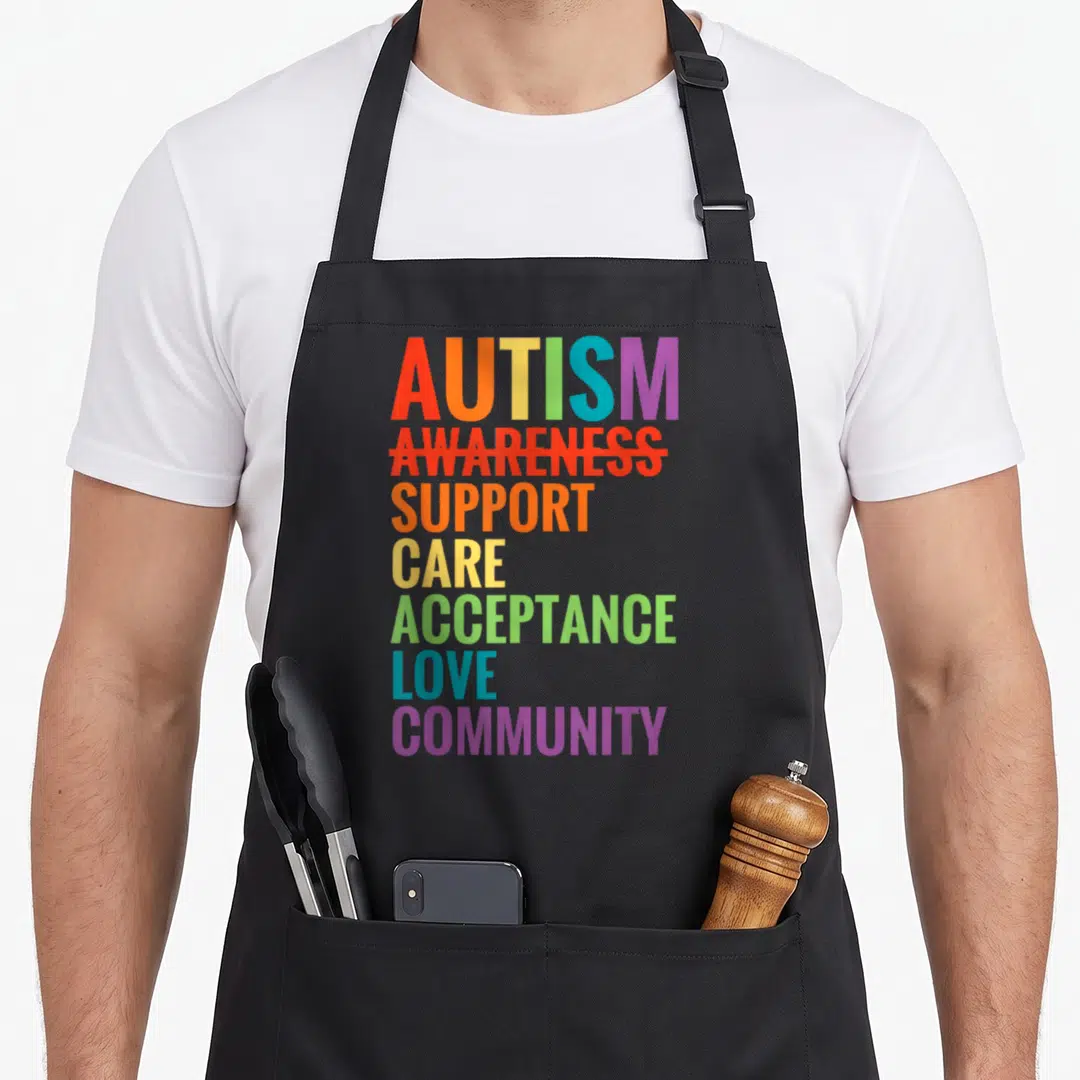 Care autism acceptance apron - Rainbow text stack reading Autism Awareness Support Care Acceptance Love Community — shown on a black cotton canvas apron against a white background