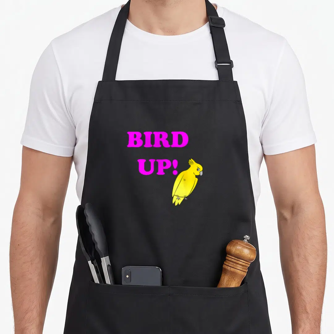 Cute bird apron - Pink BIRD UP! text stacked left of a standing yellow cartoon bird — shown on a black cotton canvas apron against a white background