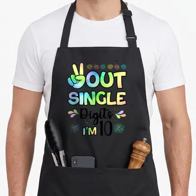Peace double digits birthday apron - Colorful peace sign hand with OUT SINGLE Digits I'M 10 text and fireworks — shown on a black cotton canvas apron against a white background