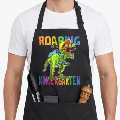 Roaring dinosaur apron - T-Rex in yellow hat and blue backpack with multicolor ROARING INTO KINDERGARTEN text — shown on a black cotton canvas apron against a white background