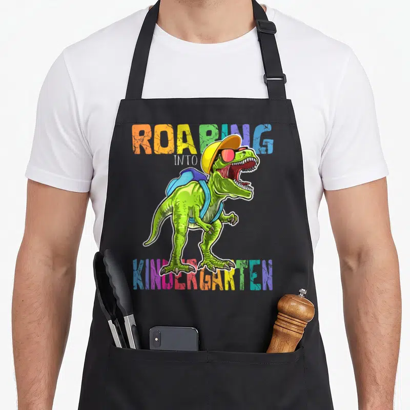 Roaring dinosaur apron - T-Rex in yellow hat and blue backpack with multicolor ROARING INTO KINDERGARTEN text — shown on a black cotton canvas apron against a white background