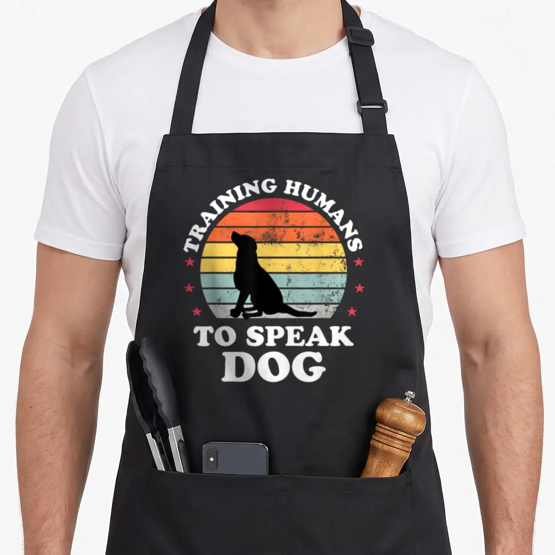 Obedience dog training apron - Black dog silhouette against red orange yellow and blue striped sun with white text — shown on a black cotton canvas apron against a white background