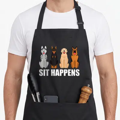 Pet dog training apron - Four cartoon dogs sitting in a row above white SIT HAPPENS text on a grey background — shown on a black cotton canvas apron against a white background