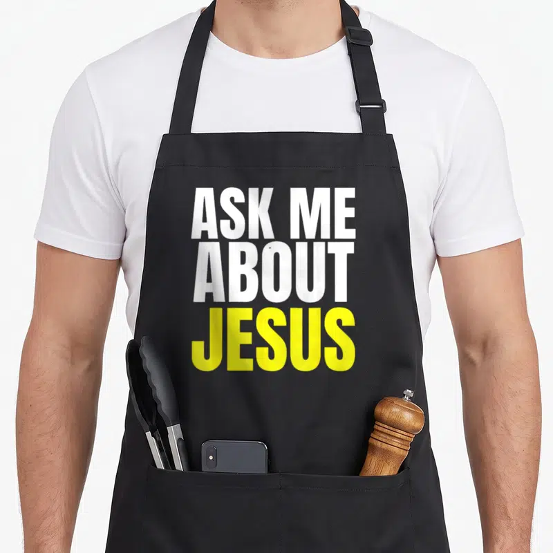 Evangelism christian apron - Stacked white and yellow text reading Ask Me About Jesus on a grey background — shown on a black cotton canvas apron against a white background