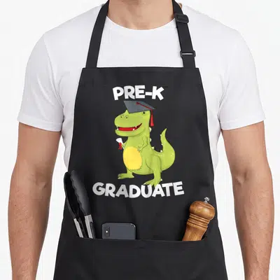 Preschool dinosaur apron - Green cartoon dinosaur wearing a grey cap and holding a white diploma with red seal — shown on a black cotton canvas apron against a white background