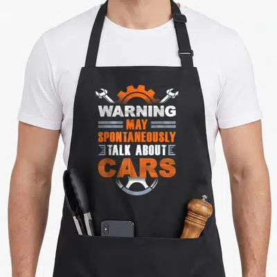 May auto mechanic apron - Distressed warning text about cars flanked by crossed wrenches and a gear above a tire base — shown on a black cotton canvas apron against a white background