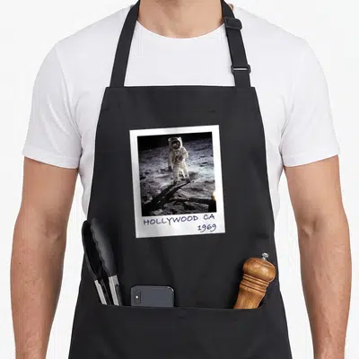 Fake moon funny apron - Astronaut in polaroid frame with handwritten HOLLYWOOD CA 1969 text below — shown on a black cotton canvas apron against a white background