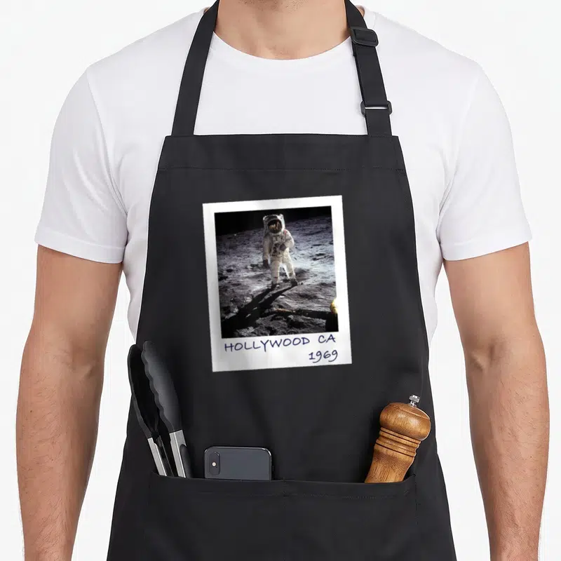 Fake moon funny apron - Astronaut in polaroid frame with handwritten HOLLYWOOD CA 1969 text below — shown on a black cotton canvas apron against a white background