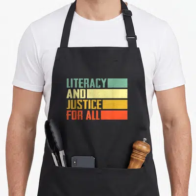 Social literacy apron - Stacked text Literacy and Justice for All beside teal yellow orange and red distressed — shown on a black cotton canvas apron against a white background
