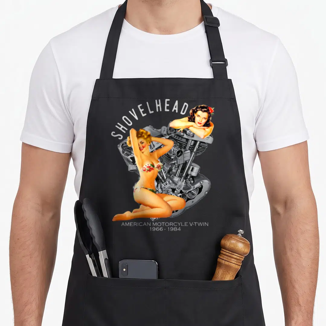 Shovelhead pinup apron - Retro pinup women posing with a grey Shovelhead V-twin engine and 1966-1984 dates — shown on a black cotton canvas apron against a white background