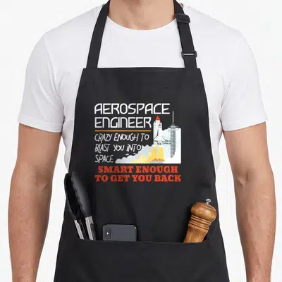 Aerospace smart apron - Cartoon space shuttle launch with AEROSPACE ENGINEER text and red yellow grey details — shown on a black cotton canvas apron against a white background