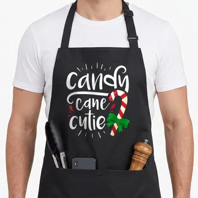 Candy womens christmas aprons - Candy cane cutie text beside a red and white striped candy with a green bow — shown on a black cotton canvas apron against a white background
