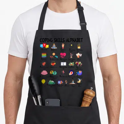 Mental tie apron - Coping Skills Alphabet grid with 27 cartoon wellness icons including yoga, journaling, and stress — shown on a black cotton canvas apron against a white background