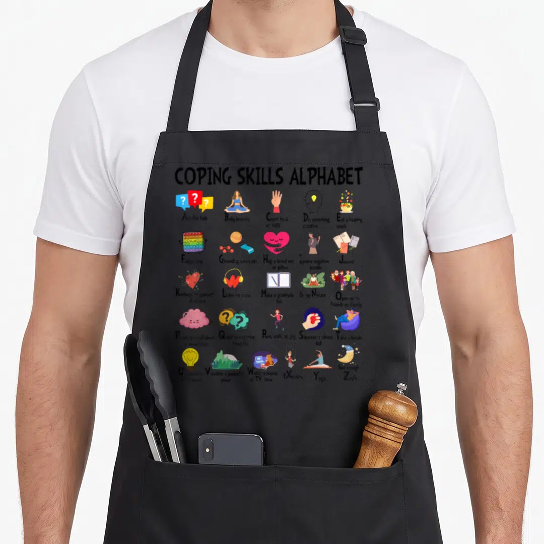 Mental tie apron - Coping Skills Alphabet grid with 27 cartoon wellness icons including yoga, journaling, and stress — shown on a black cotton canvas apron against a white background