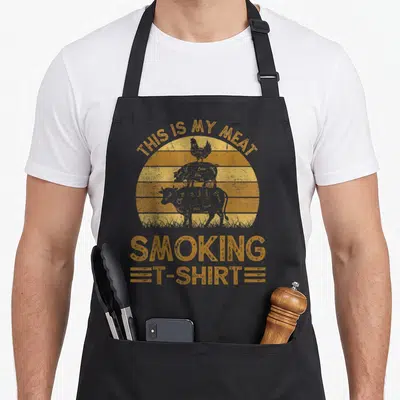 Smoking meat aprons - Orange distressed sun with stacked chicken pig cow silhouettes and THIS IS MY MEAT SMOKING — shown on a black cotton canvas apron against a white background