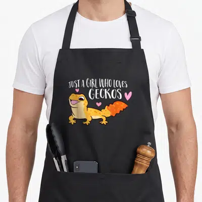 Gecko lizard leopard apron - Cartoon orange gecko with pink hearts under arched white text reading JUST A GIRL WHO — shown on a black cotton canvas apron against a white background