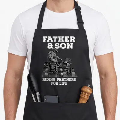 Riding father son apron - FATHER & SON RIDING PARTNERS FOR LIFE text above father and son on ATVs holding hands — shown on a black cotton canvas apron against a white background