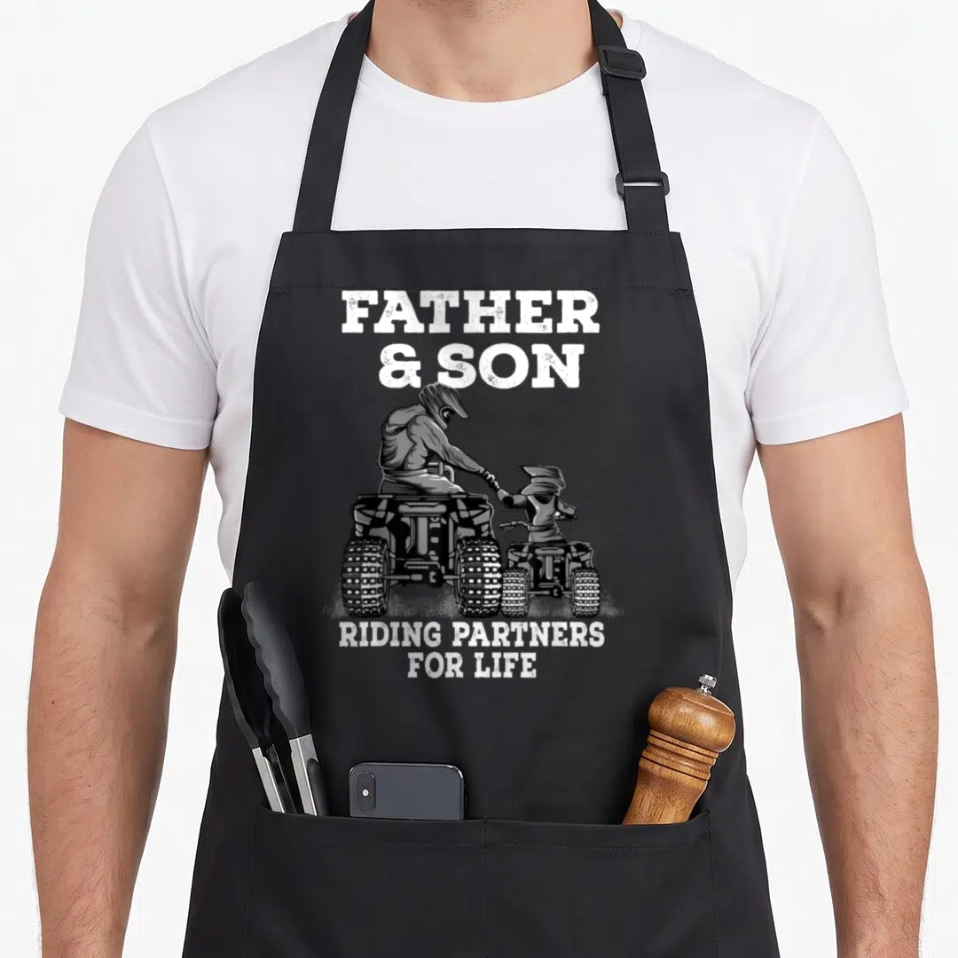 Riding father son apron - FATHER & SON RIDING PARTNERS FOR LIFE text above father and son on ATVs holding hands — shown on a black cotton canvas apron against a white background