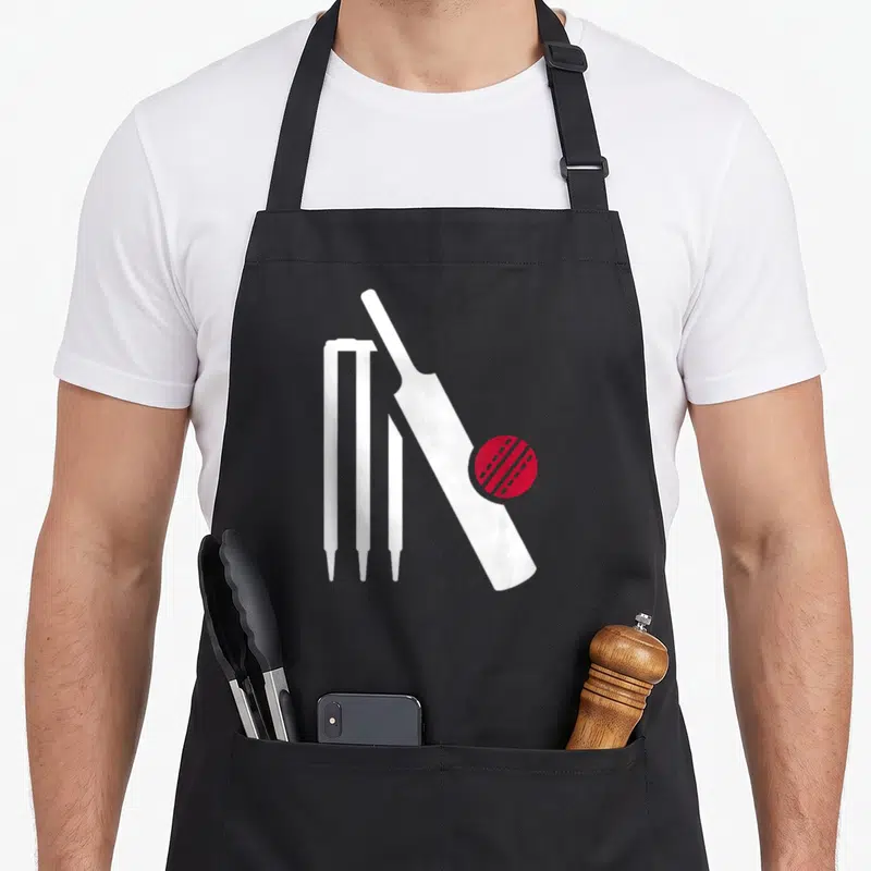 Bat cricket apron - White bat leaning on three stumps with a red ball graphic — shown on a black cotton canvas apron against a white background