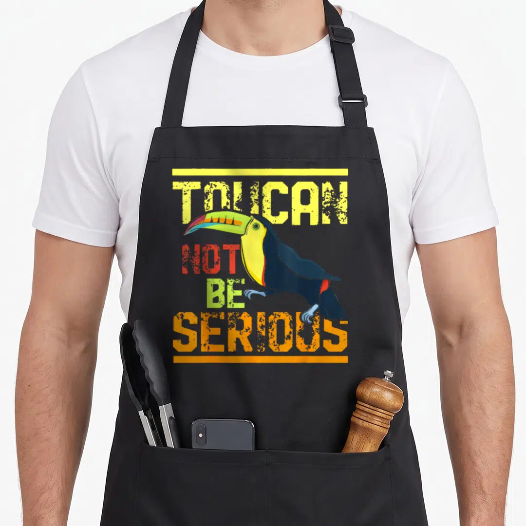 Zoo birds apron - Cartoon toucan with multicolored beak overlapping stacked text TOUCAN NOT BE SERIOUS — shown on a black cotton canvas apron against a white background