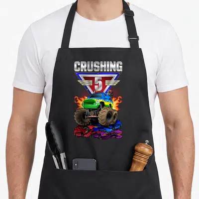 Crushing 5th truck apron - Blue monster truck crushing three cars below silver CRUSHING 5 text and triangle logo — shown on a black cotton canvas apron against a white background