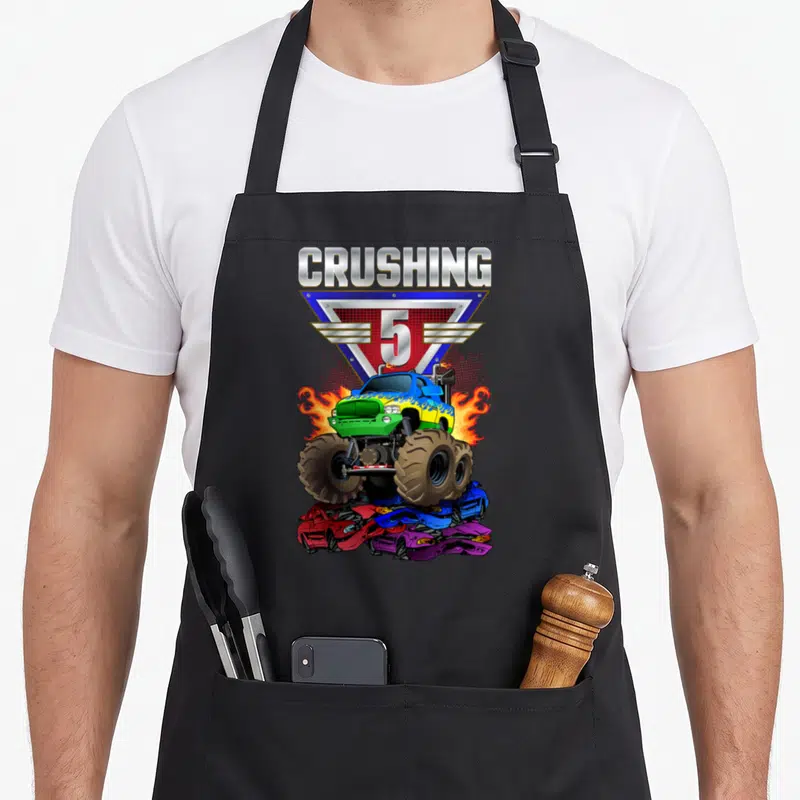 Crushing 5th truck apron - Blue monster truck crushing three cars below silver CRUSHING 5 text and triangle logo — shown on a black cotton canvas apron against a white background