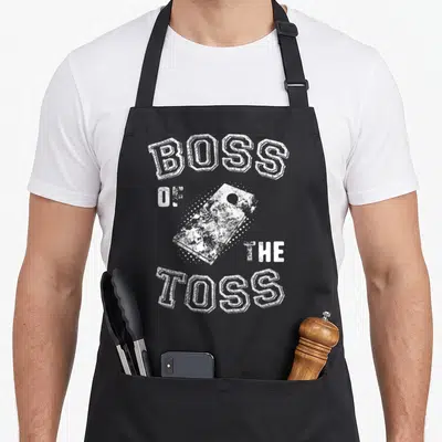 Cornhole distressed apron - White distressed bean bag surrounded by BOSS OF THE TOSS block letters — shown on a black cotton canvas apron against a white background