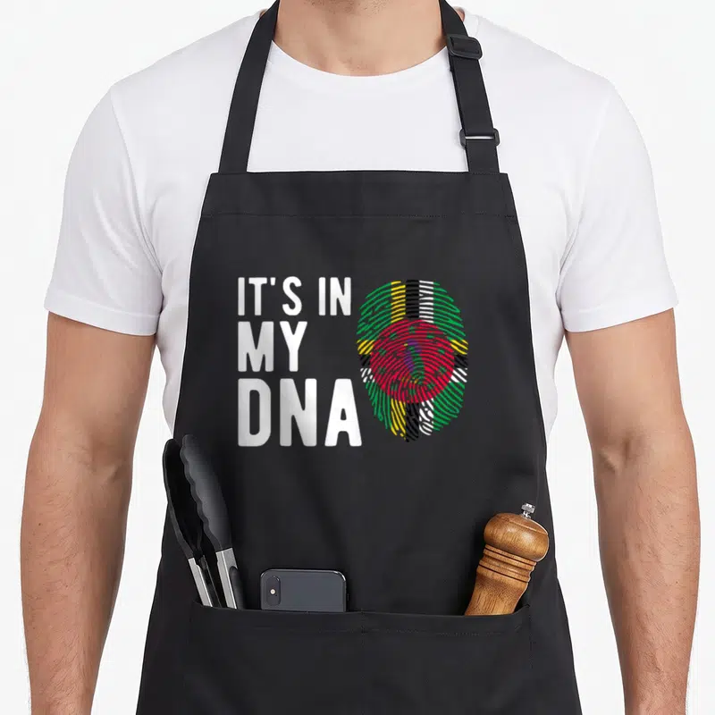 Dna dominica apron - White text IT'S IN MY DNA beside fingerprint with red blue green yellow and white flag colors — shown on a black cotton canvas apron against a white background