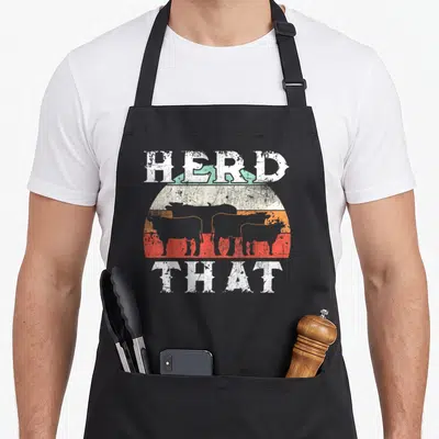 Herd farmer apron - Distressed HERD THAT text with cattle silhouettes over orange and red sunset bands — shown on a black cotton canvas apron against a white background