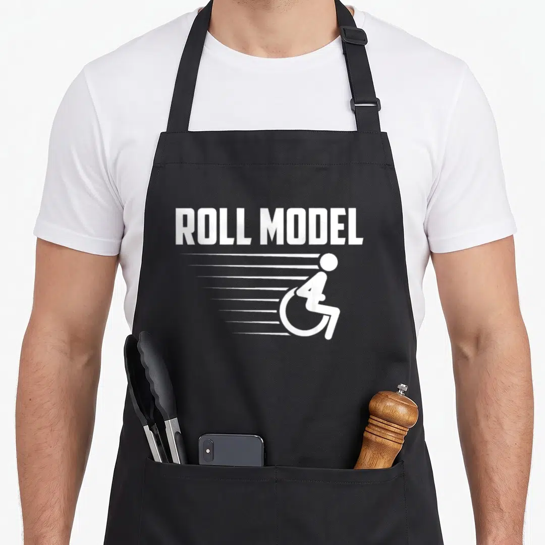 Model wheelchair apron - ROLL MODEL text above white minimalist person in wheelchair with horizontal speed lines — shown on a black cotton canvas apron against a white background