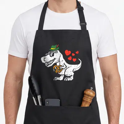Rex oktoberfest apron - Cartoon T-Rex in green hat holding orange pretzel with four red hearts — shown on a black cotton canvas apron against a white background