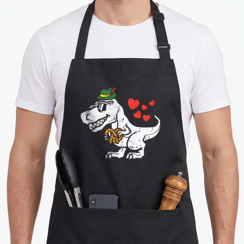 Rex oktoberfest apron - Cartoon T-Rex in green hat holding orange pretzel with four red hearts — shown on a black cotton canvas apron against a white background