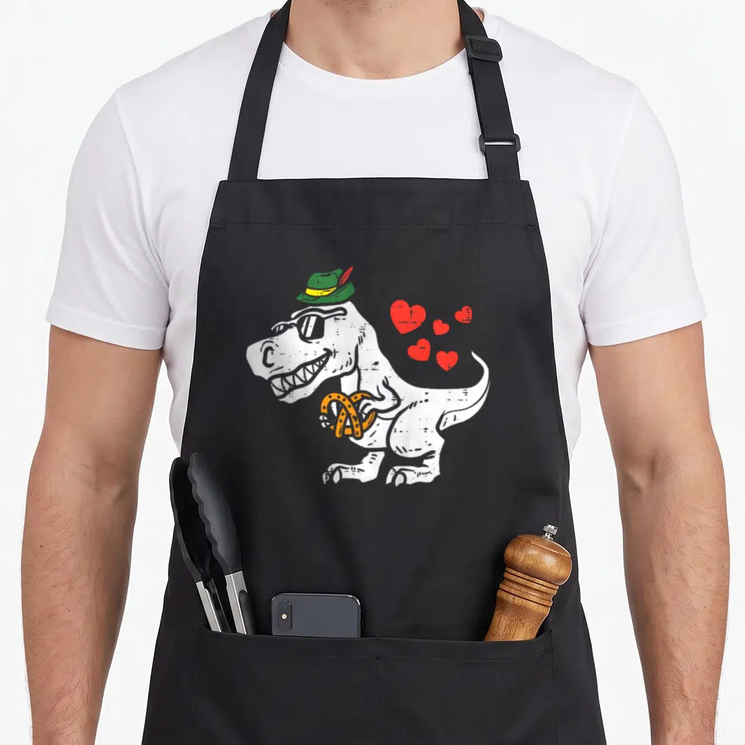Rex oktoberfest apron - Cartoon T-Rex in green hat holding orange pretzel with four red hearts — shown on a black cotton canvas apron against a white background