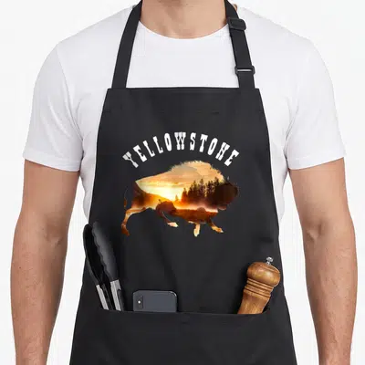 Funny national apron - Yellowstone text arched over bison silhouette filled with orange sunset landscape and trees — shown on a black cotton canvas apron against a white background