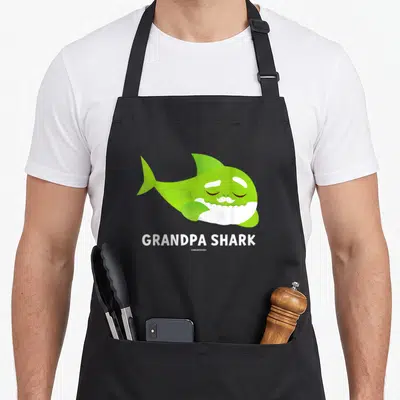 Pinkfong shark grandpa apron - Green cartoon shark with white mustache and GRANDPA SHARK text — shown on a black cotton canvas apron against a white background