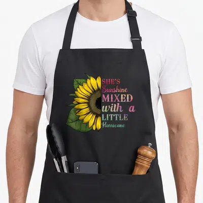 She sunflower apron - Realistic sunflower with rainbow text SHE'S Sunshine MIXED with a LITTLE Hurricane — shown on a black cotton canvas apron against a white background