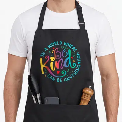 Kind floral apron - Colorful Be Kind typography surrounded by a quote circle with red hearts and green leaves — shown on a black cotton canvas apron against a white background
