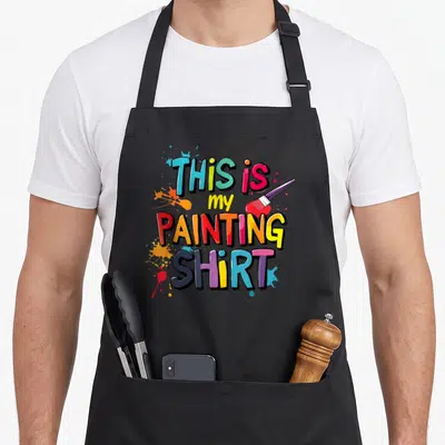 Present painting apron - Colorful text THIS IS my PAINTING SHIRT with paint splatters and brush — shown on a black cotton canvas apron against a white background