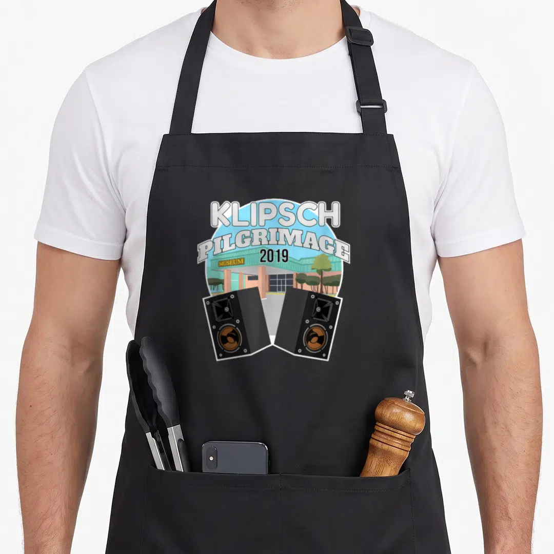 Hifi audiophile apron - Cartoon Klipsch Pilgrimage 2019 museum building with two black speakers and green trees — shown on a black cotton canvas apron against a white background
