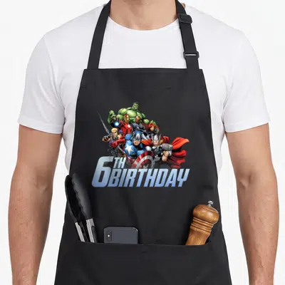 Shot 6th birthday apron - Avengers cartoon group clustered over 6TH BIRTHDAY text in blue red green white — shown on a black cotton canvas apron against a white background