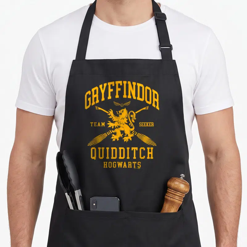 Quidditch arch apron - Yellow GRYFFINDOR text arching over a lion and crossed broomsticks — shown on a black cotton canvas apron against a white background